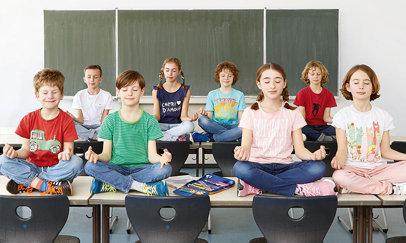 Kinder sitzen im Schneidersitz vor einer Tafel auf den Schultischen